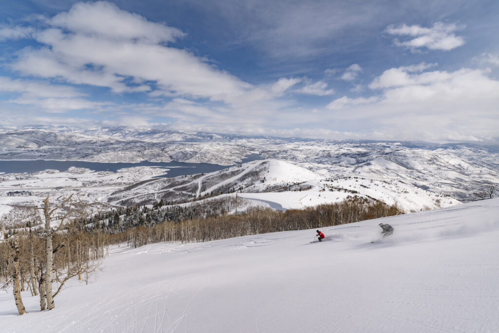 Two skiers ski down Deer Valley’s new expanded terrain overlooking the shimmering Jordanelle Reservoir and the Wasatch Mountains.