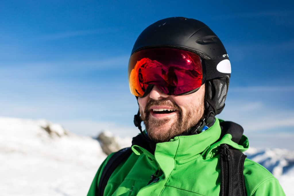 Close-up of happy male skier with large oversized ski goggles standing on the top of a mountain ready for skiing.