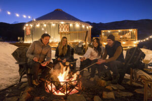Group of friends gathered around fire at dusk with yurt behind them